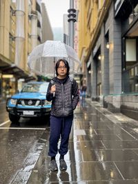 Portrait of young asian man with umbrella walking in the rain on city street against buildings