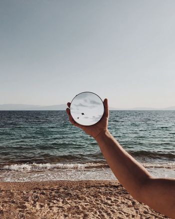 Cropped hand of person holding mirror at beach against sky