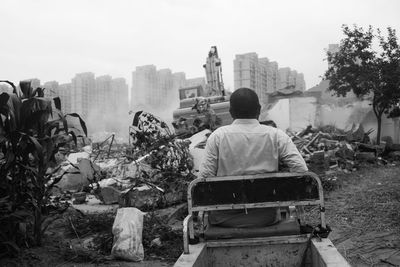 Rear view of man on seat against bulldozer demolishing houses