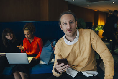 Portrait of confident businessman holding smart phone while sitting against female colleagues at office in cafeteria