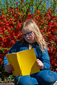 Young school age girl reading from yellow textbook in park against bright red flowers background.
