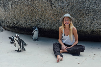 Portrait of smiling young woman sitting by penguins at beach