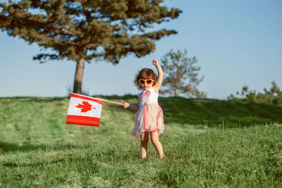 Cute girl holding canadian flag while standing on grass against trees and sky