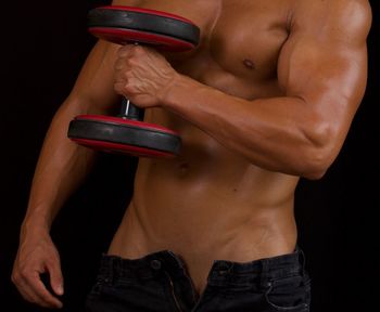 Midsection of shirtless man exercising with dumbbell against black background