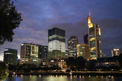 Illuminated buildings by river against sky at dusk