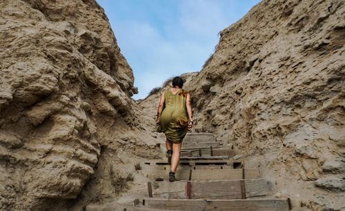 Rear view of woman moving up on steps amidst rock formation against sky