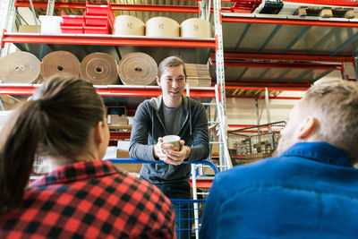 Manual worker having coffee while discussing with male and female colleagues in warehouse