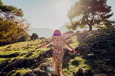 Back view of carefree female in summer dress standing on rocky hill in highlands on sunny day