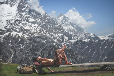 Shirtless man relaxing on bench against snowcapped mountain against sky