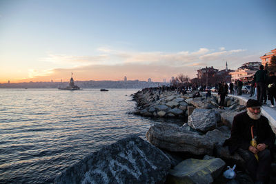 People on rocks by sea against sky during sunset