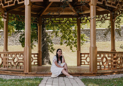 Portrait of beautiful young woman wearing white dress sitting under wooden pavilion in park.