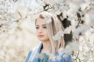 Portrait of happy young blonde female in blue jacket embracing in white flowers