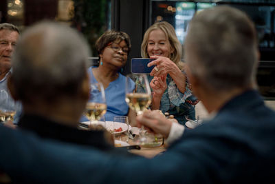 Smiling senior women photographing male friends holding wineglasses in restaurant