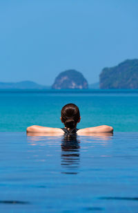  young woman wearing bikini standing in blue infinity swimming pool looking at the view of ocean 