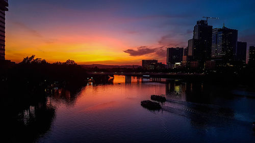 Silhouette buildings by river against sky during sunset