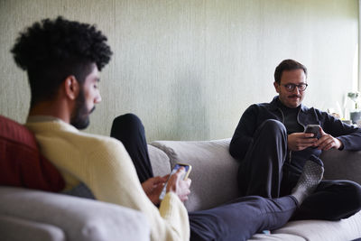 Gay couple relaxing on sofa at home and using phones