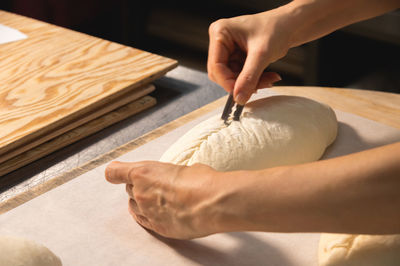 Close-up of a baker's female hands cutting a loaf of dough into a loaf of bread with a blade before