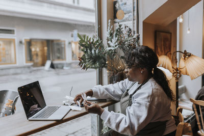 Side view of mature cafe owner with bills and laptop sitting at table by glass window in coffee shop