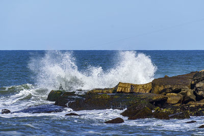 Waves splashing on rocks against clear sky