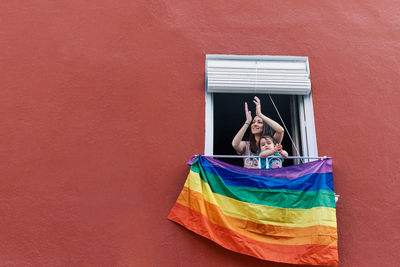 Young woman in a mask clapping from the window with the gay flag on a red background