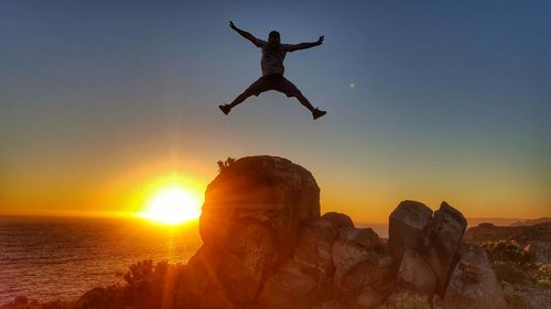 Silhouette man jumping on rock against sea during sunset