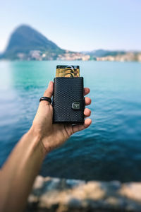 Close-up of hand holding camera by sea against sky