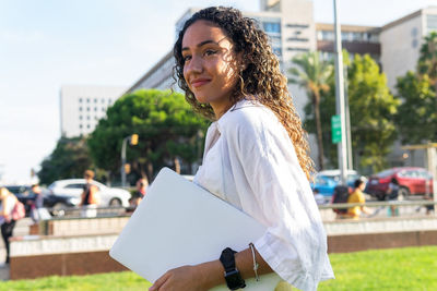 Side view of hispanic young woman in white shirt with curly hair carrying laptop and looking away while spending time in park on sunny summer day in city