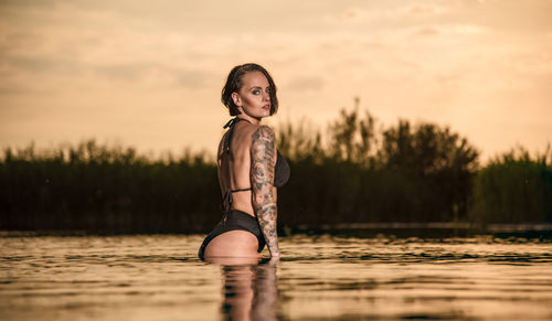 Portrait of young woman standing in lake against sky during sunset