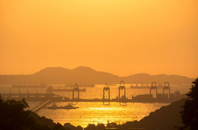 Silhouette cranes at dock in sea against orange sky