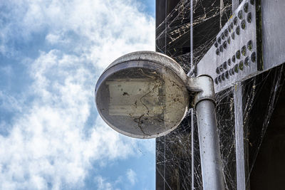 Spider web on a public street lamp attached to a steel railway bridge, with a blue sky with white cl