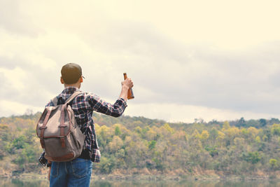 Rear view of man with backpack holding bottle while standing by lake against sky