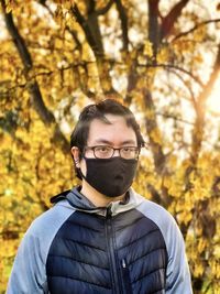 Young asian man in eyeglasses and face mask standing against flowering laburnum tree at sunset.