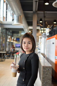 Portrait of smiling young woman holding coffee while standing in restaurant