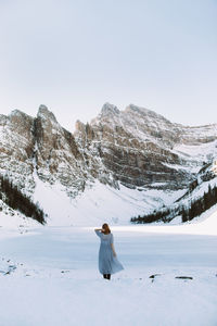 Back view of anonymous woman in dress touching hair and standing near frozen lake louise against snowy mountain range on winter day in alberta, canada