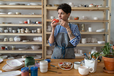 Thoughtful creative young woman ceramist drinking tea and resting at workplace in pottery studio