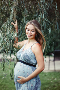 Smiling pregnant woman touching abdomen while standing on grass