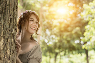 Smiling young woman standing by tree trunk in forest