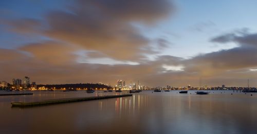 Illuminated city by sea against sky at sunset