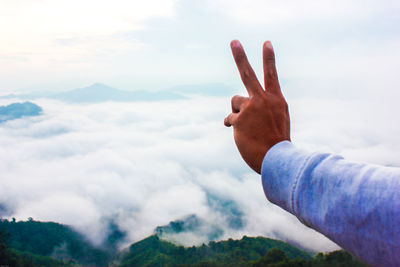 Cropped hand gesturing peace sign against cloudy sky