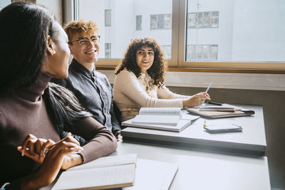 Smiling young multiracial friends sitting together at desk in university classroom