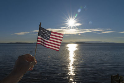 Cropped hand of man holding american flag over sea against sky during sunny day
