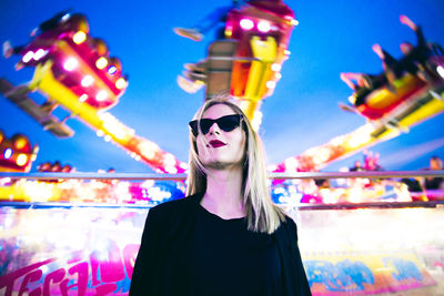 Low angle view of woman wearing sunglasses while standing against illuminated ride