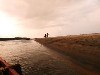 People walking on shore at beach against sky during sunset