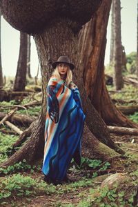 Young woman wrapped in blanket standing against tree trunk in forest