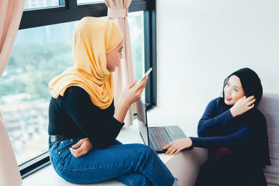 Young woman using mobile phone while sitting on window