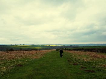 Mid distance of man and dog walking on grassy field against cloudy sky
