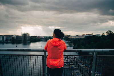 Rear view of female athlete in raincoat looking at city while standing on footbridge over sea during sunset