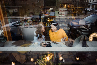 Female bloggers with camera and laptop sitting at cafe seen through window glass