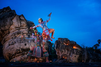 People in traditional clothing performing on rock formation against blue sky at dusk