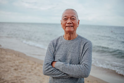 Portrait of senior man with arms crossed standing at beach against sky during sunset